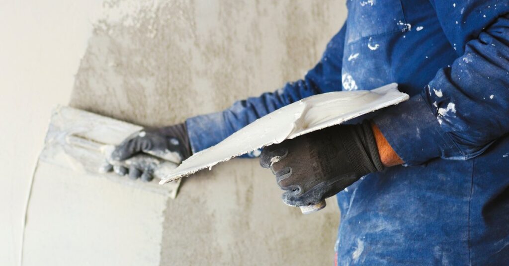 A worker is wearing a dirty painters shirt while they use a plastering tool to spread stucco on a wall.