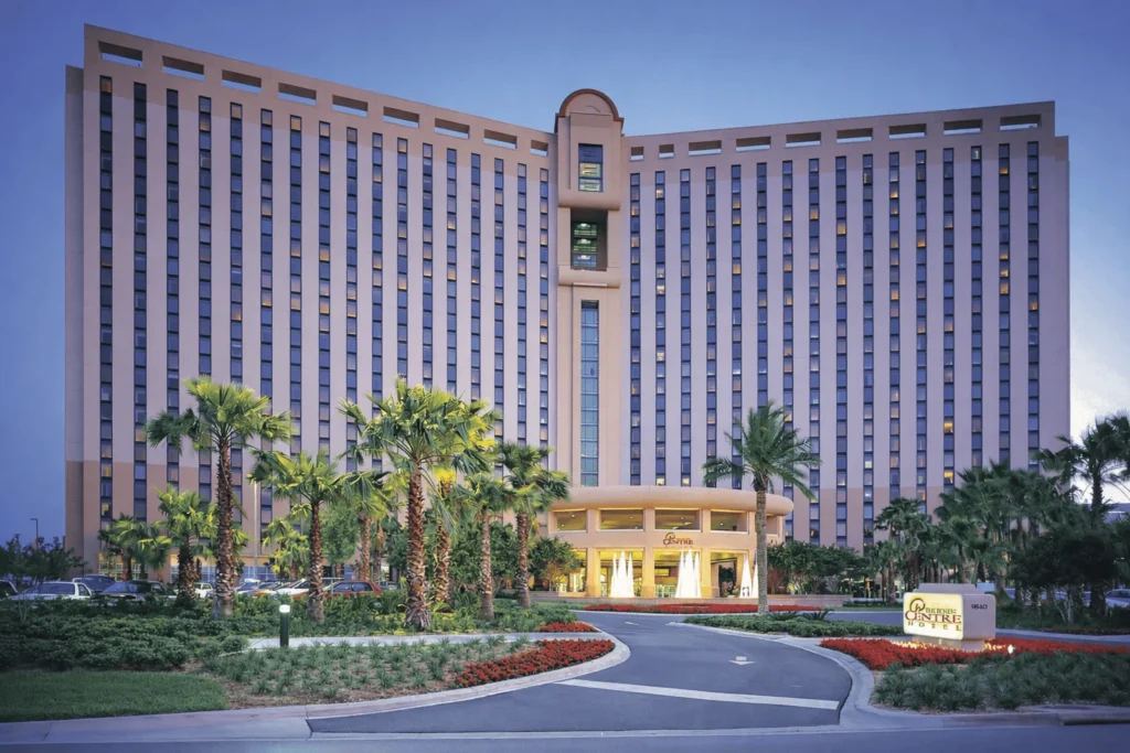 A tall, modern hotel building with many windows, palm trees, and neatly landscaped gardens at the entrance. The curved driveway leads to a well-lit main entrance at dusk. A sign reads The Point Hotel & Suites.
