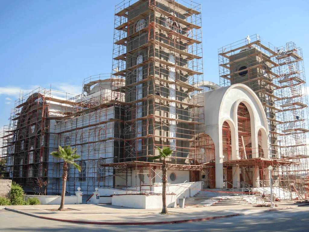 A large building with arched entrances and towers is surrounded by scaffolding, indicating ongoing construction or renovation. Two palm trees are in front, and the sky is clear and blue.