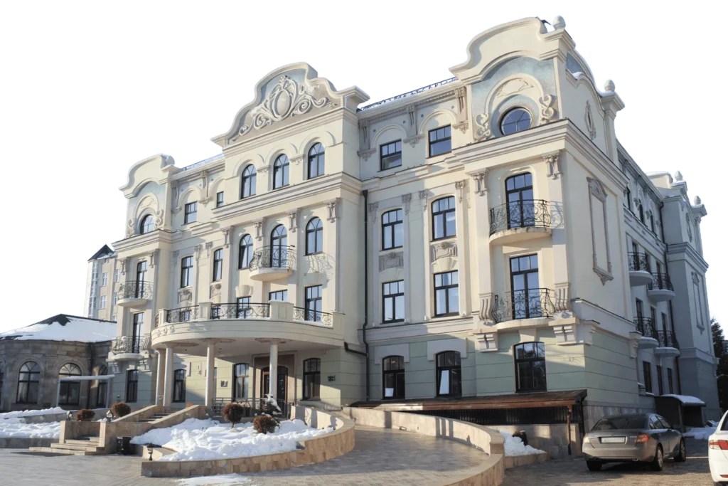 Elegant, ornate mansion with three stories, arched windows, decorative balconies, and detailed trim. Snow covers the ground, and two cars are parked in front on a curved driveway.