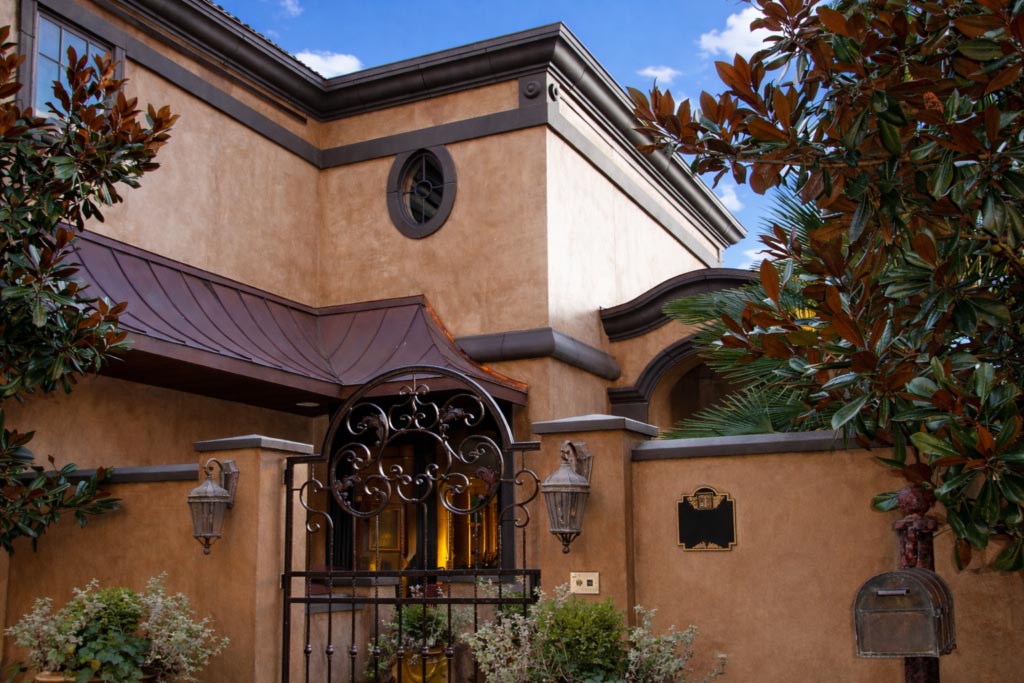 A tan stucco house with dark trim, an ornate metal gate, lantern-style lights, and trees framing the entrance under a blue sky with clouds.
