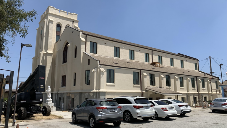 A large, light-colored building with tall windows and a tower stands in bright sunlight. Several cars are parked in front, and construction equipment is nearby, indicating renovation work.