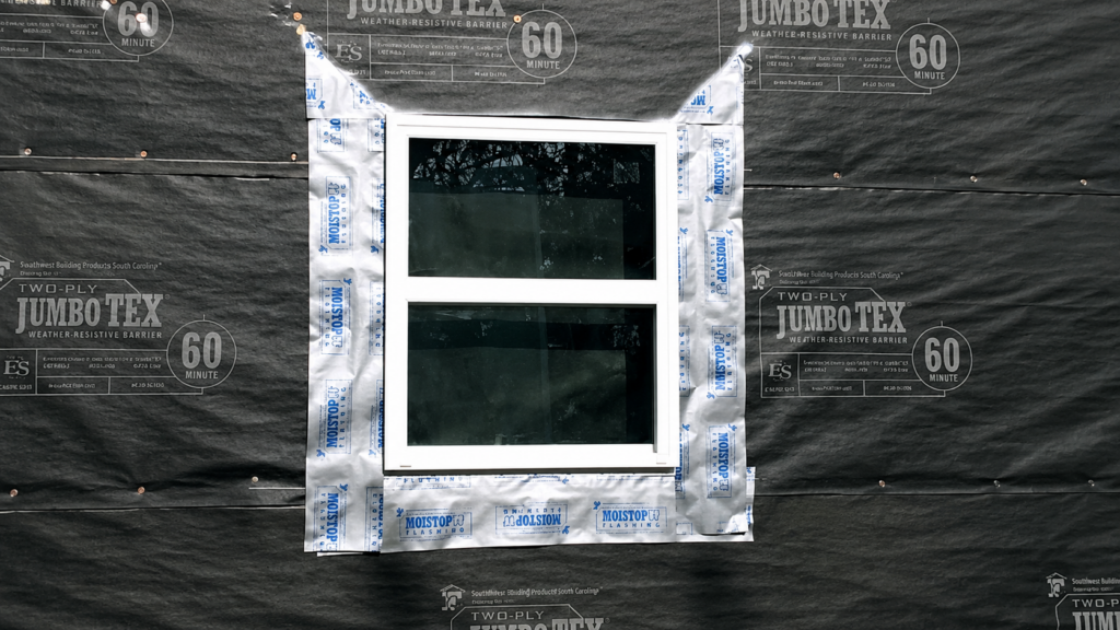 A white-framed window is installed in a wall covered with black building wrap. The window is surrounded by silver flashing tape for waterproofing, and trees are reflected in the window glass.